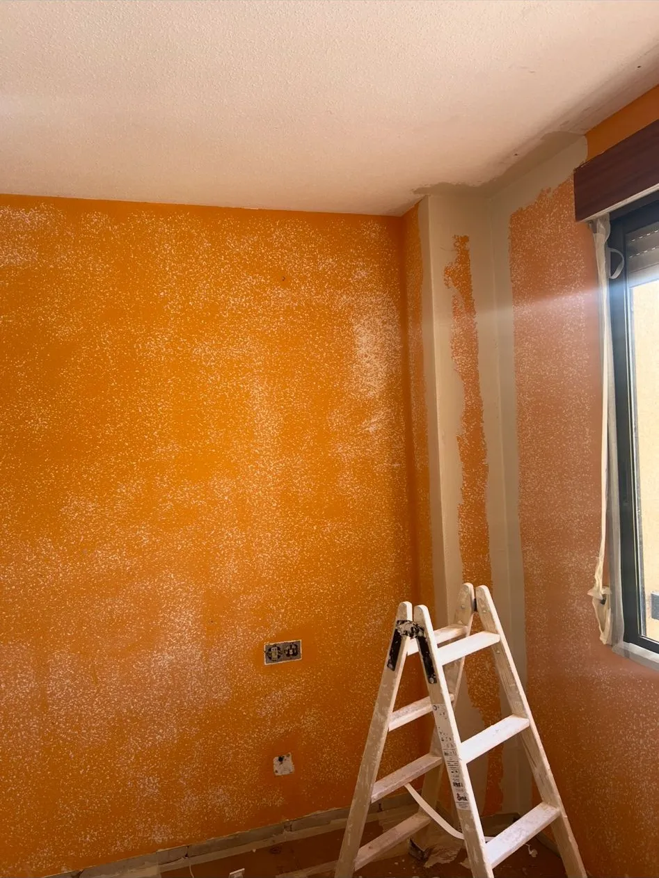 Esquina de un dormitorio con paredes naranjas con gotelé, una escalera de pintor apoyada y el suelo sin pavimentar