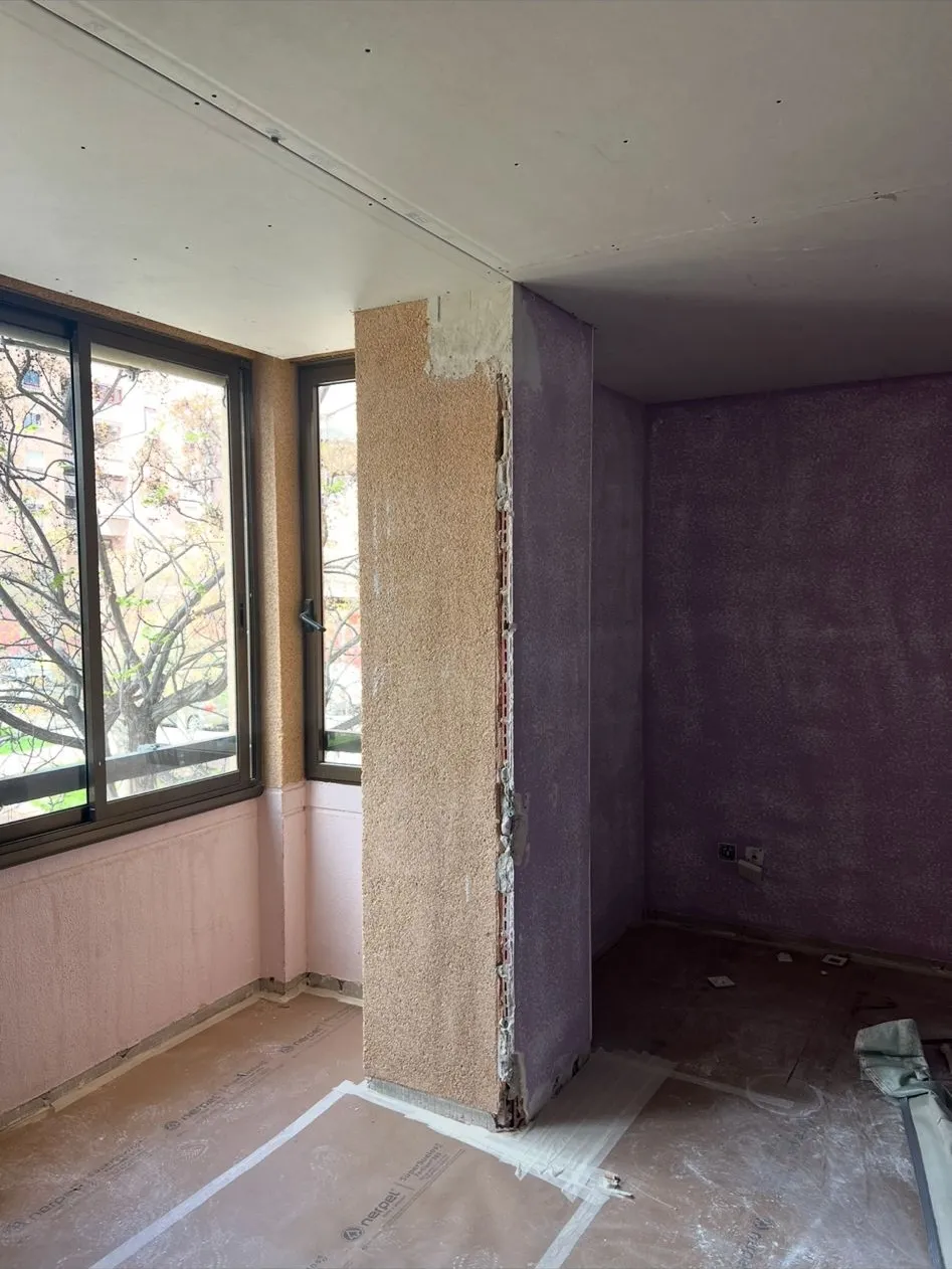 Estancia en obra con ventanal en esquina, pilar con gotelé beige, paredes lilas con gotelé y zócalo rosa bajo la ventana